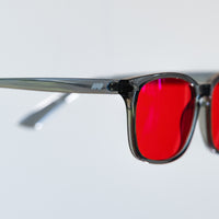 Close-up of a pair of square-frame sunglasses with translucent dark gray frames and bright red tinted lenses, shown against a light gray background.
