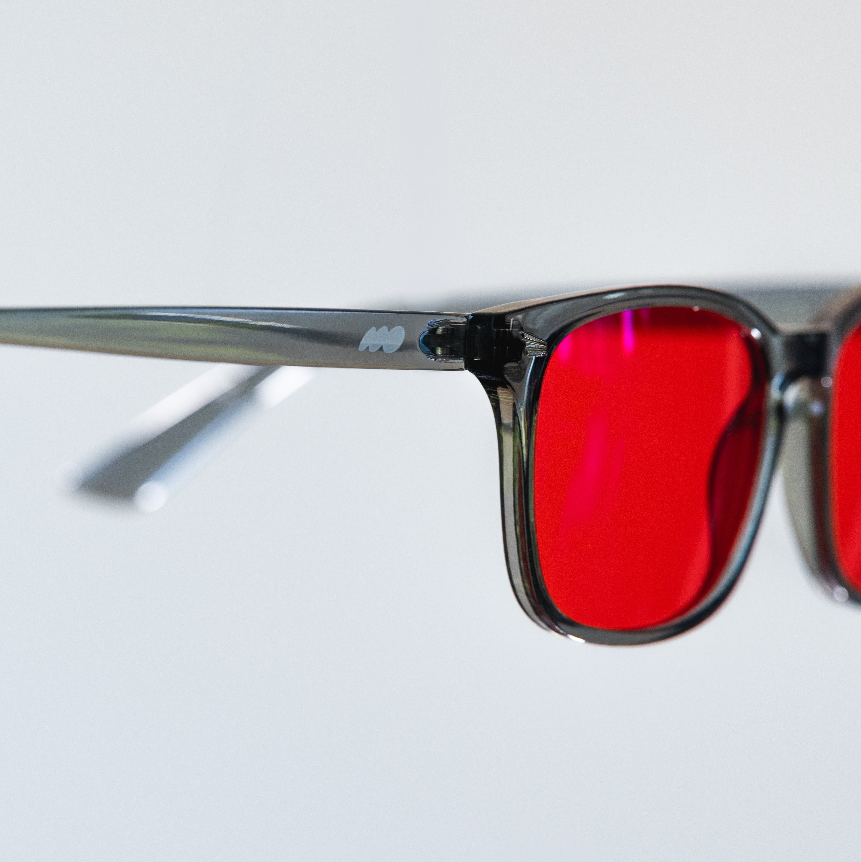 Close-up of a pair of square-frame sunglasses with translucent dark gray frames and bright red tinted lenses, shown against a light gray background.