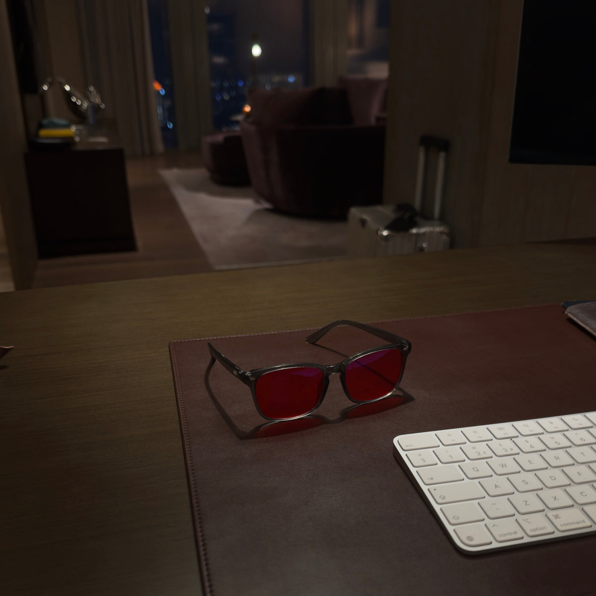 Blue light glasses with red lenses resting on a brown leather desk mat next to a white keyboard on a wooden desk, with a blurred modern living room in the background.